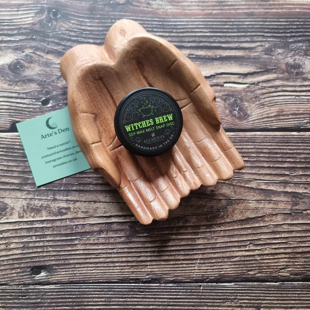 A gothic-themed wax melt tin labelled Witches Brew, displayed in a wooden pair of hands on a wooden surface next to a green Arte's Den business card 