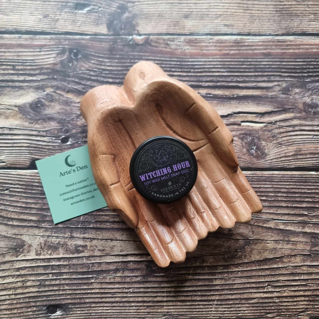 A gothic-themed wax melt tin labelled Witching Hour, displayed in a wooden pair of hands on a wooden surface next to a green Arte's Den business card 