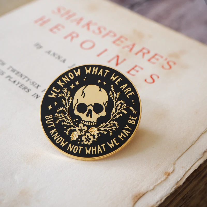 Pin with skull design and text on a book which reads "we know what we are but not what we may be"
