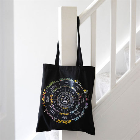 Black tote bag with colourful wheel of the year design hanging on a white banister. 