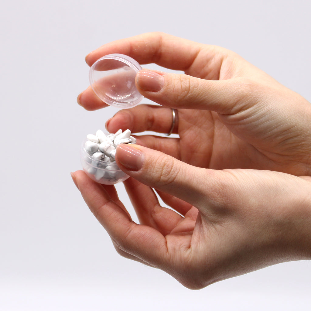 A pair of hands opening a see-through capsule holding a white jasper bracelet 
