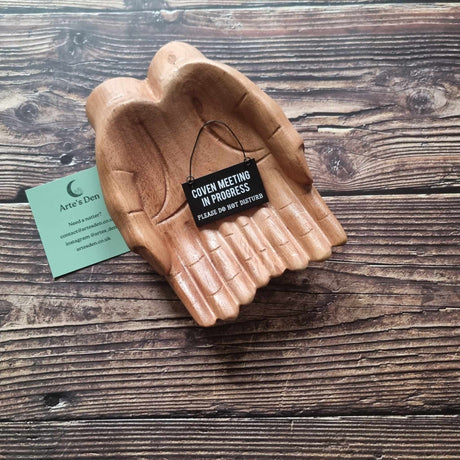 A pair of wooden hands hold a black mini sign with the phrase 'Coven meeting in progress please do not disturb' placed on a wooden surface next to a green Arte's Den business card. 
