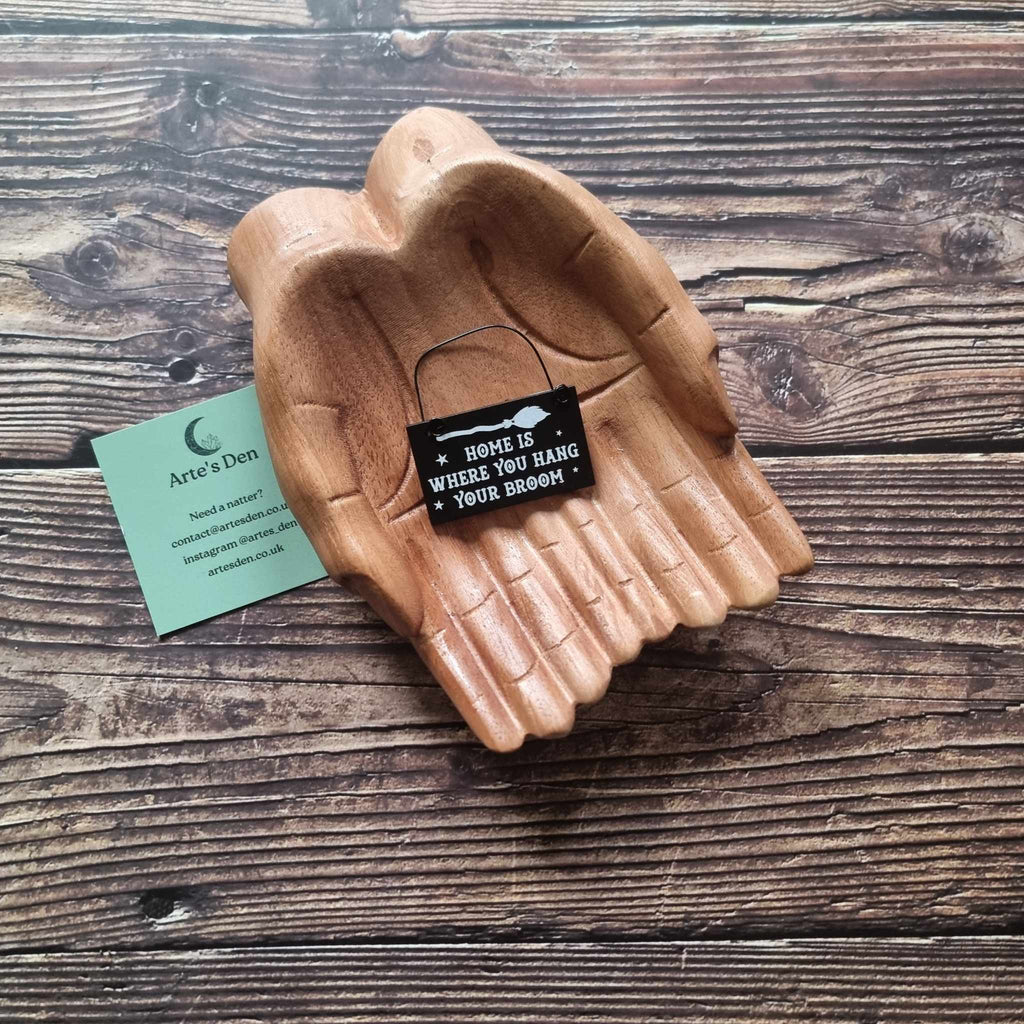 A pair of wooden hands hold a black mini sign with the phrase 'Home is where you have your broom' placed on a wooden surface next to a green Arte's Den business card. 