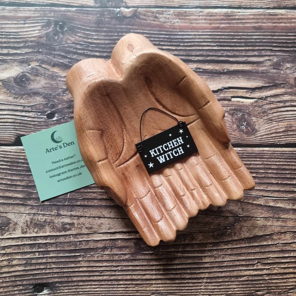 A pair of wooden hands hold a black mini sign with the phrase 'Kitchen Witch' placed on a wooden surface next to a green Arte's Den business card. 