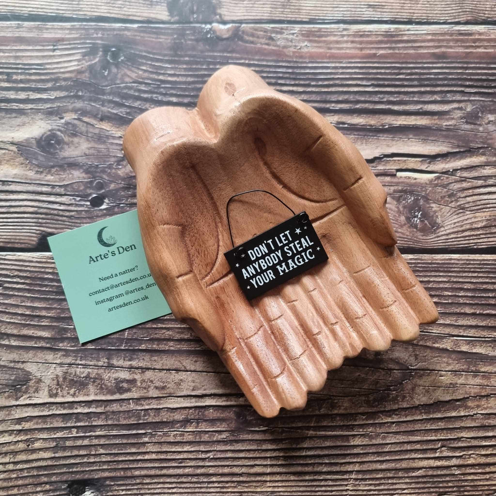 A pair of wooden hands hold a black mini sign with the phrase 'Don't let anybody steal your magic' placed on a wooden surface next to a green Arte's Den business card. 