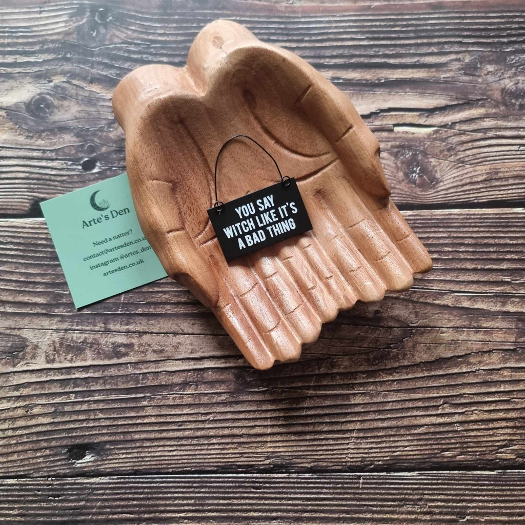 A pair of wooden hands hold a black mini sign with the phrase 'You say witch like it's a bad thing' placed on a wooden surface next to a green Arte's Den business card. 