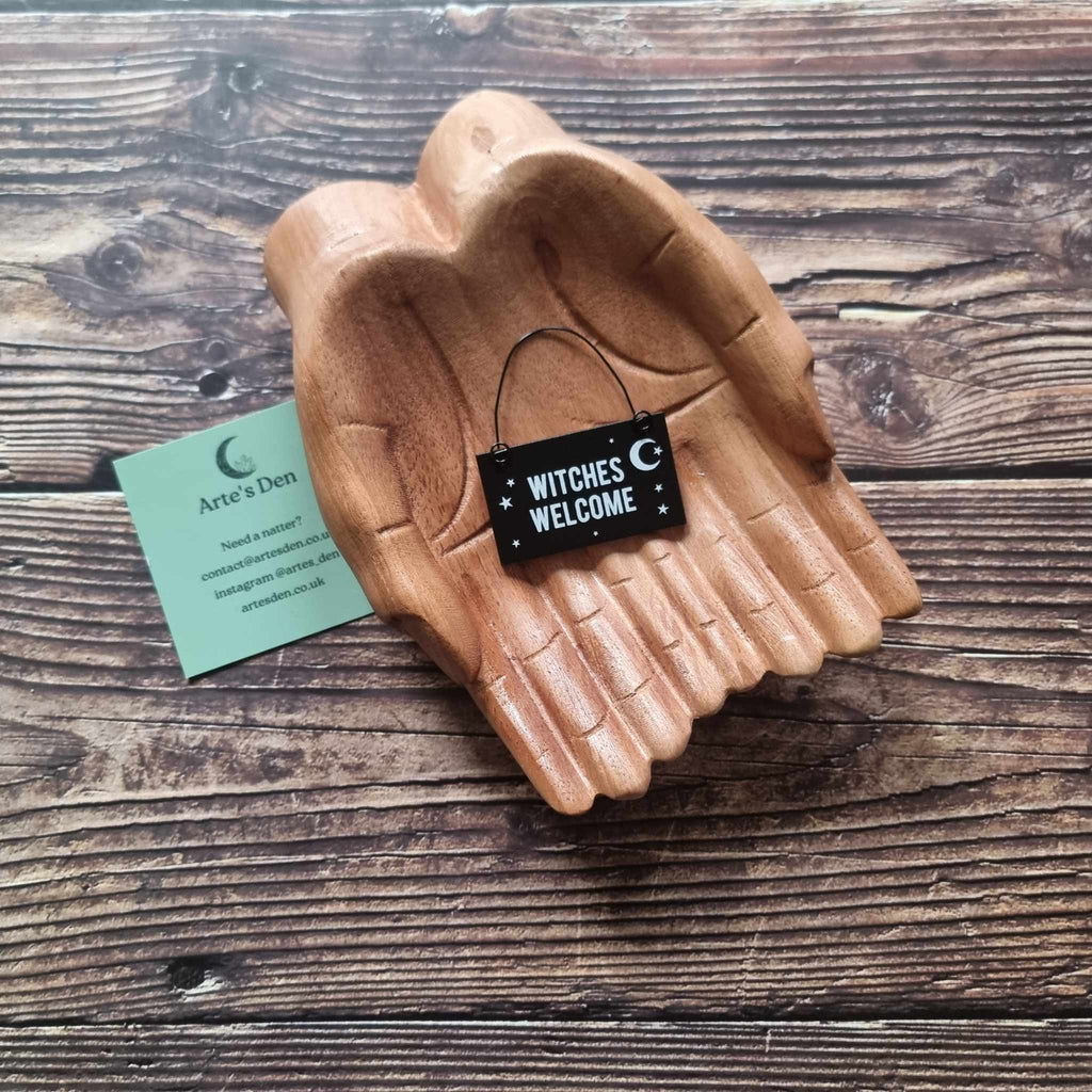 A pair of wooden hands hold a black mini sign with the phrase 'Witches Welcome' placed on a wooden surface next to a green Arte's Den business card. 