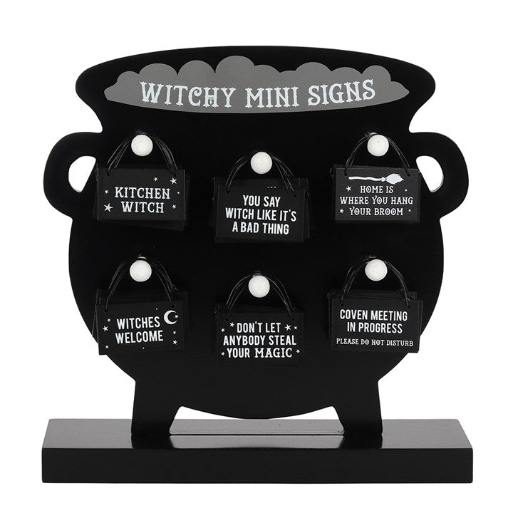 A collection of black mini witch signs with white font displayed against a black cauldron holder with white hanging pins states "witchy mini signs"