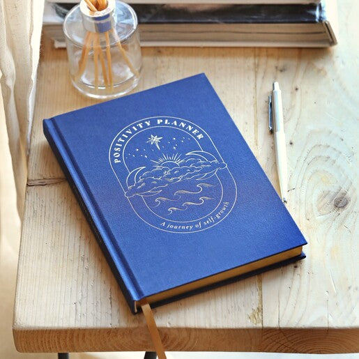 A navy blue journal with a gold detail on the cover, featuring "Positivity Planner" with a gold page edge.