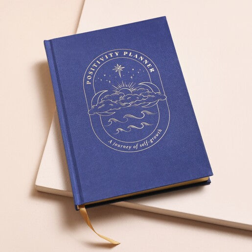 A navy blue journal with a gold detail on the cover, featuring "Positivity Planner" and moon/cloud designs with a gold page edge.