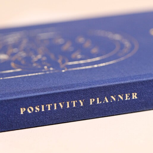 A navy blue journal with a gold detail on the cover, featuring "Positivity Planner" on the spine.