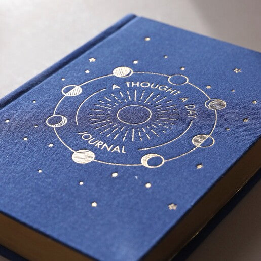 A navy blue journal with "A thought a day journal" written on the front with a moon phase design. 