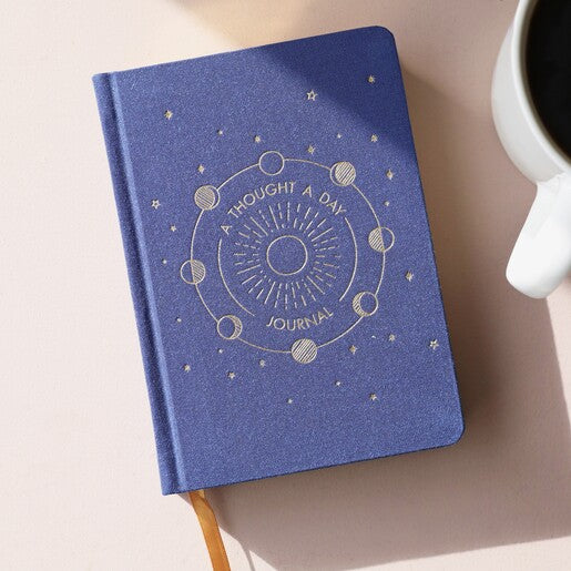 A navy blue journal with "A thought a day journal" written on the front with a moon phase design. 