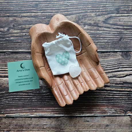 A clear quartz crystal heart stone with a small drawstring pouch, placed on a wooden pair of hands.