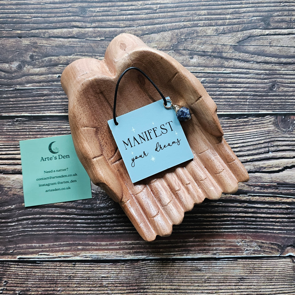 A mini sign with attached crystal charm, inscribed with the phrase "Manifest your dreams" displayed on a pair of wooden hands on a wooden surface. 