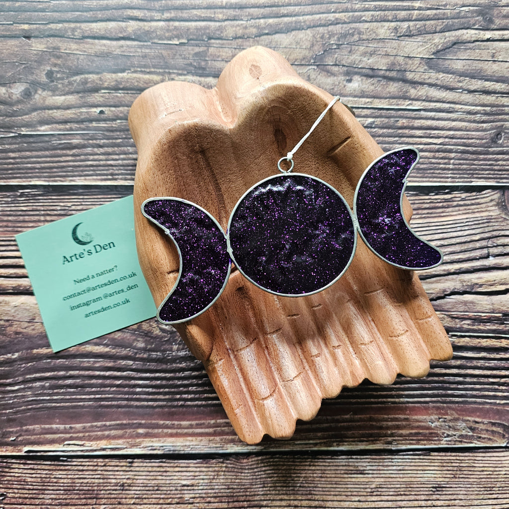 A purple sparkly triple moon suncatcher on a pair of wooden hands. 