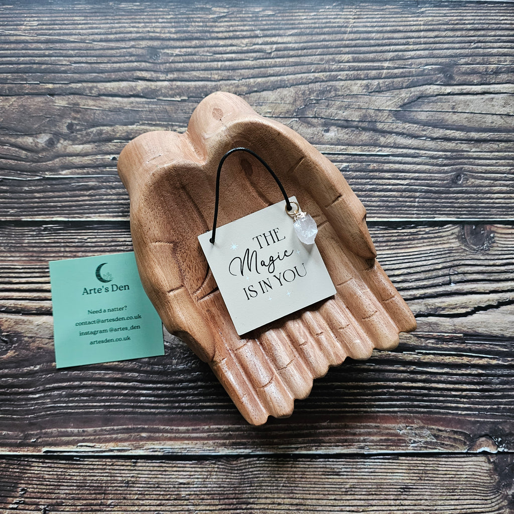 A mini sign with attached crystal charm, inscribed with the phrase "The magic is in you" displayed on a pair of wooden hands on a wooden surface. 