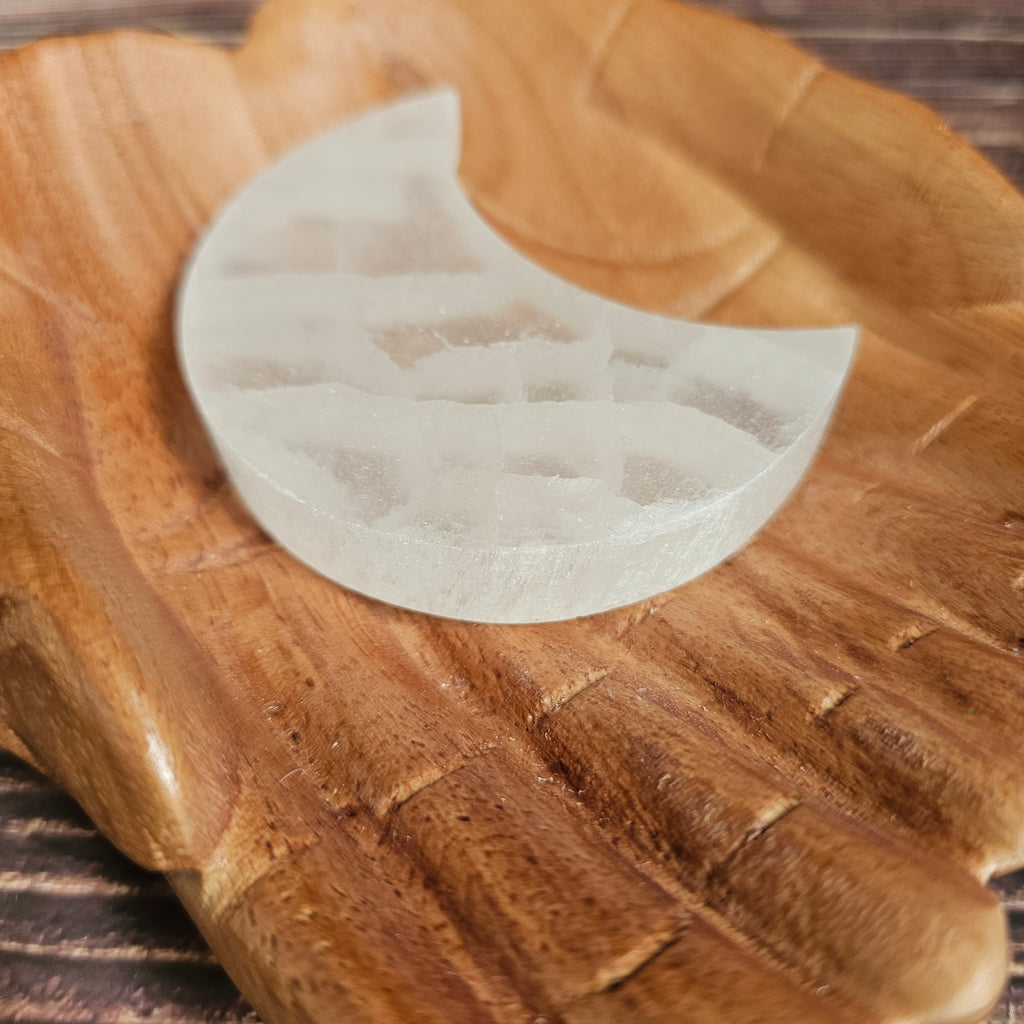 A selenite crystal charging plate shaped as a crescent moon, placed on a wooden pair of hands