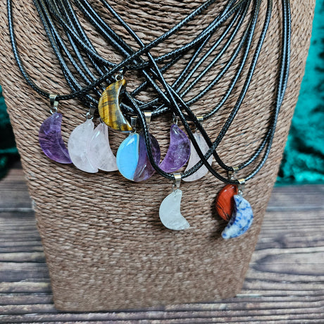 A collection of crescent moon shaped crystal pendants in various colours, displayed on a woven mannequin, with black cord necklaces.