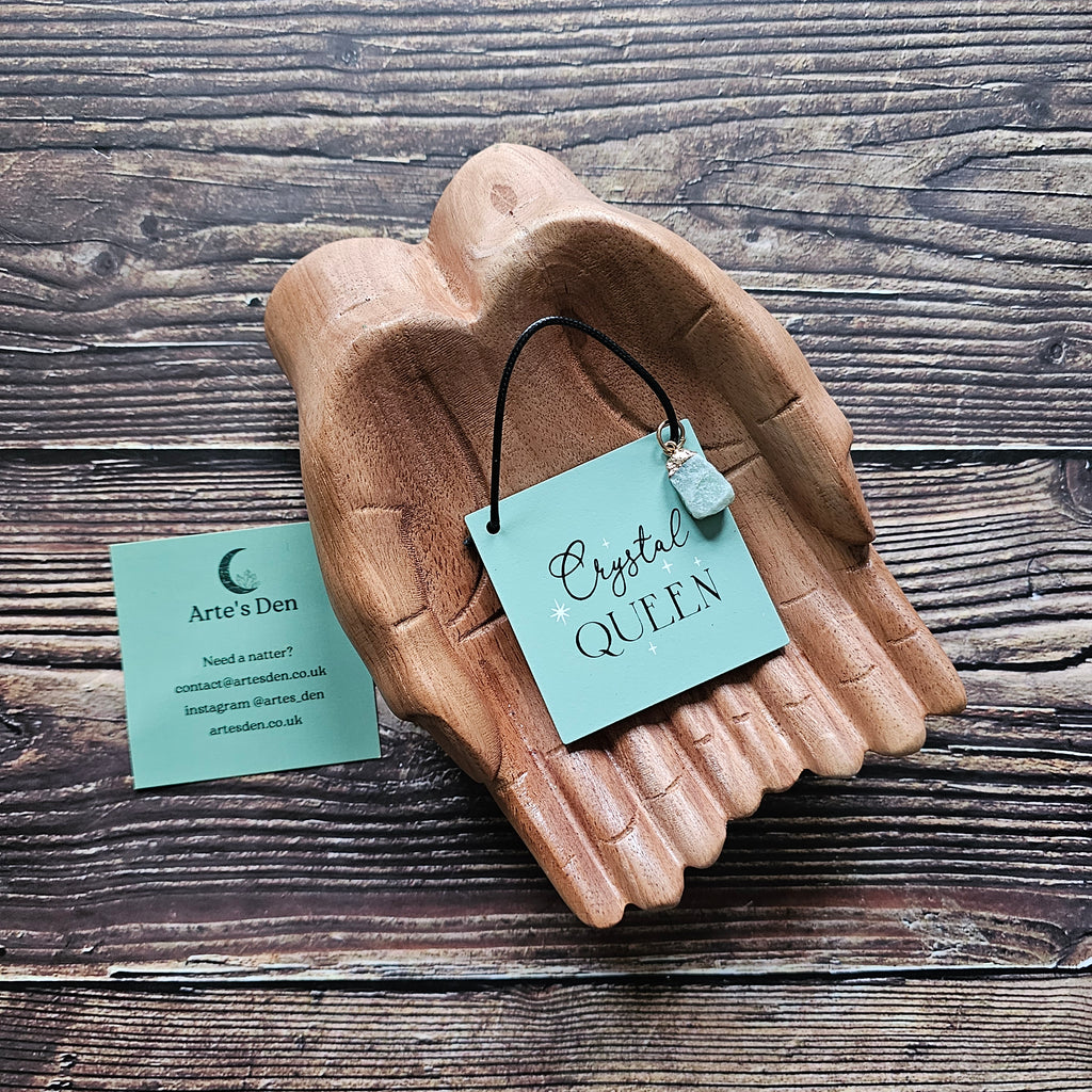 A mini sign with attached crystal charm, inscribed with the phrase "Crystal Queen" displayed on a pair of wooden hands on a wooden surface. 