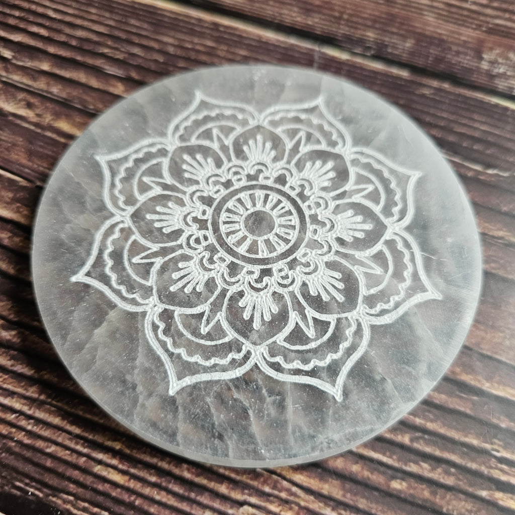 A round selenite charging plate with a mandala pattern etched into it, placed on a wooden surface.