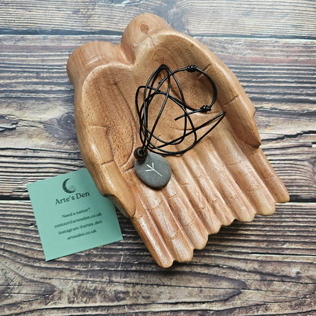 A hand-carved stone pendant with a Algiz rune attached to a black cord, placed on a wooden pair of hands on a wooden surface. A business card is also visible in the foreground.