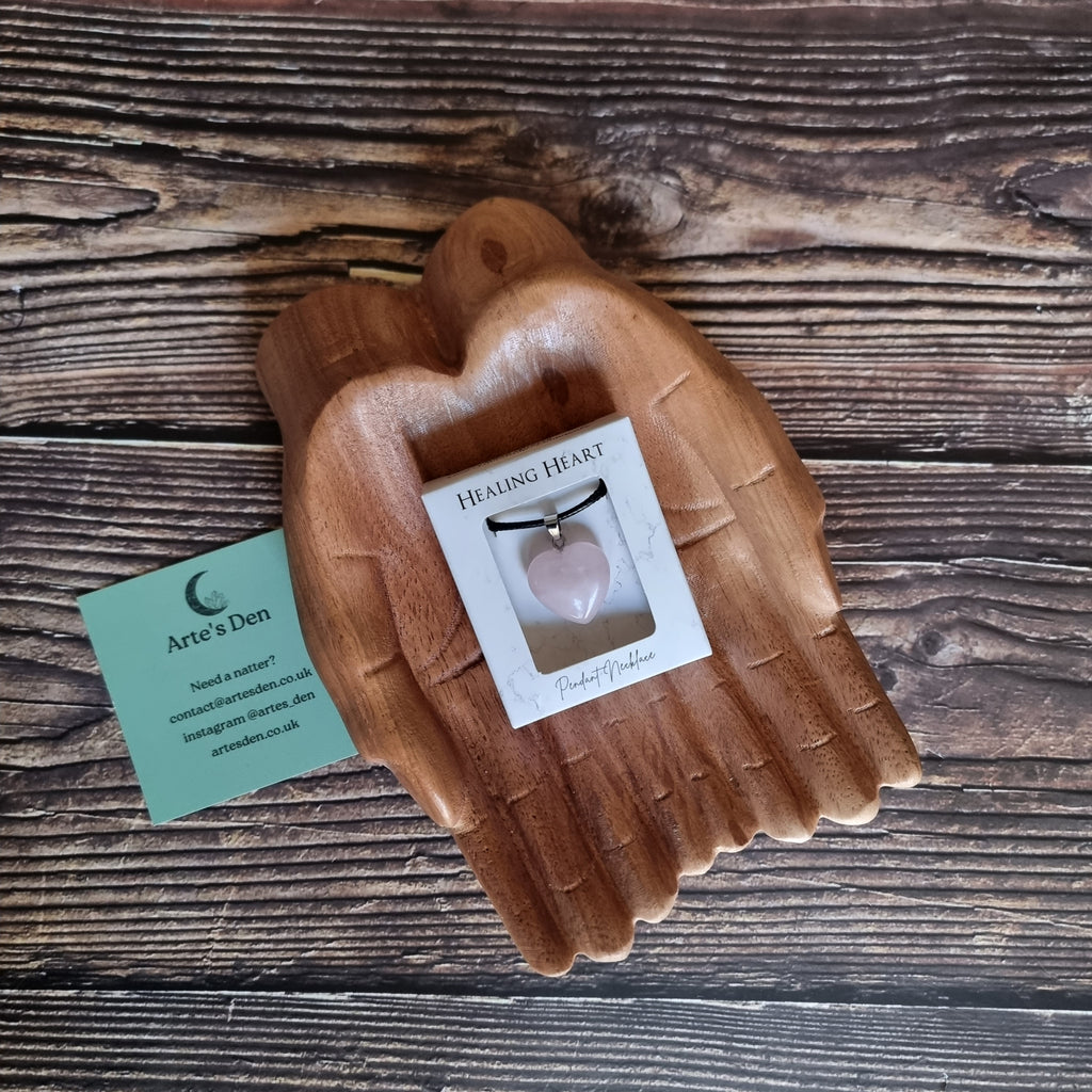 A pink crystal heart on a black cord necklace inside a white display box, displayed on a pair of wooden hands