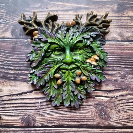 A small face-shaped plaque featuring a green man design with leaf patterns, acorns, and toadstools against a wooden background