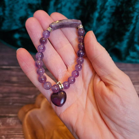 A hand holding a purple amethyst bead bracelet with a heart-shaped bead and bar quartz accents.