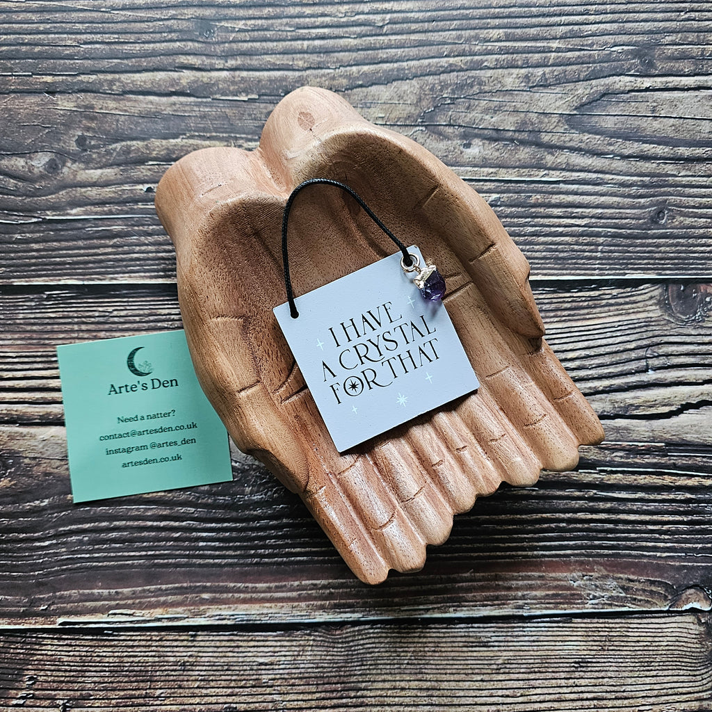 A mini sign with attached crystal charm, inscribed with the phrase "I Have a Crystal For That" displayed on a pair of wooden hands on a wooden surface. 