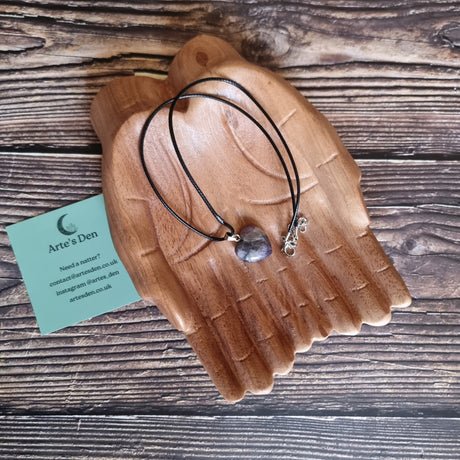 A purple crystal heart on a black cord necklace displayed on a pair of wooden hands
