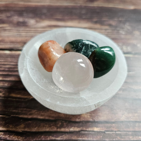 A white selenite bowl on a wooden surface, with several small colorful stones arranged on it.