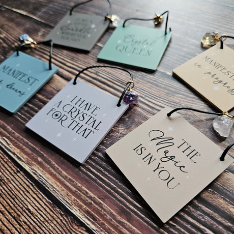 A selection of mini signs with attached crystal charms, each inscribed with different phrases, displayed on a wooden surface.