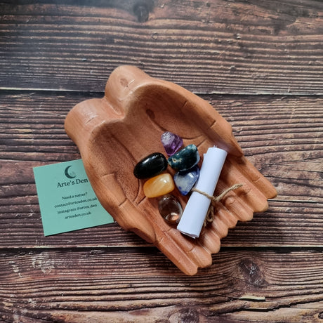 A collection of polished crystals and a scroll of paper displayed in a pair of wooden hands, with a business card that reads 'Arte's Den'.