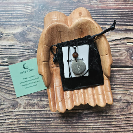 A hand-carved Fehu rune stone pendant with a black cord, presented on a wooden surface with a business card beside it.