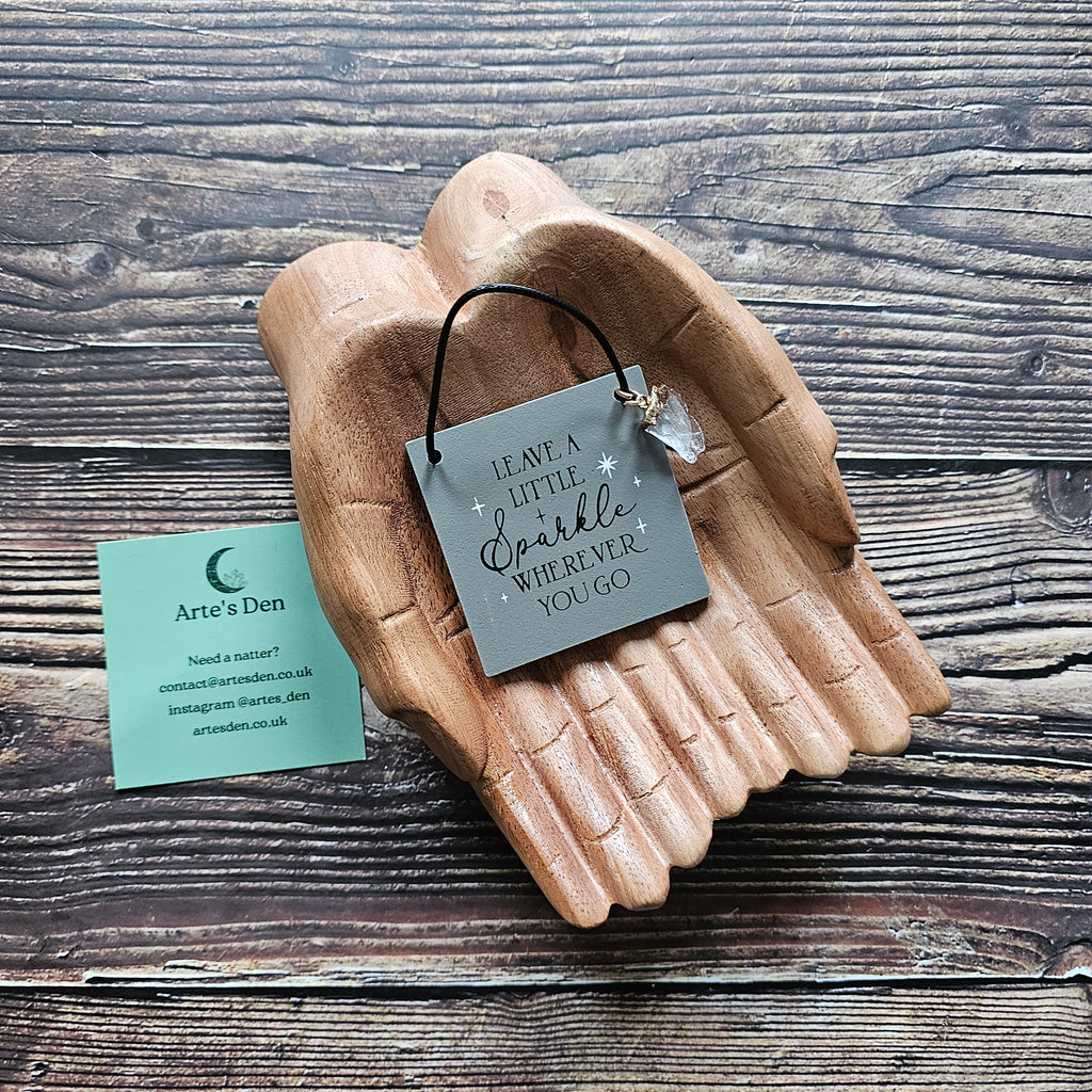 A mini sign with attached crystal charm, inscribed with the phrase "Leave a little sparkle wherever you go" displayed on a pair of wooden hands on a wooden surface. 