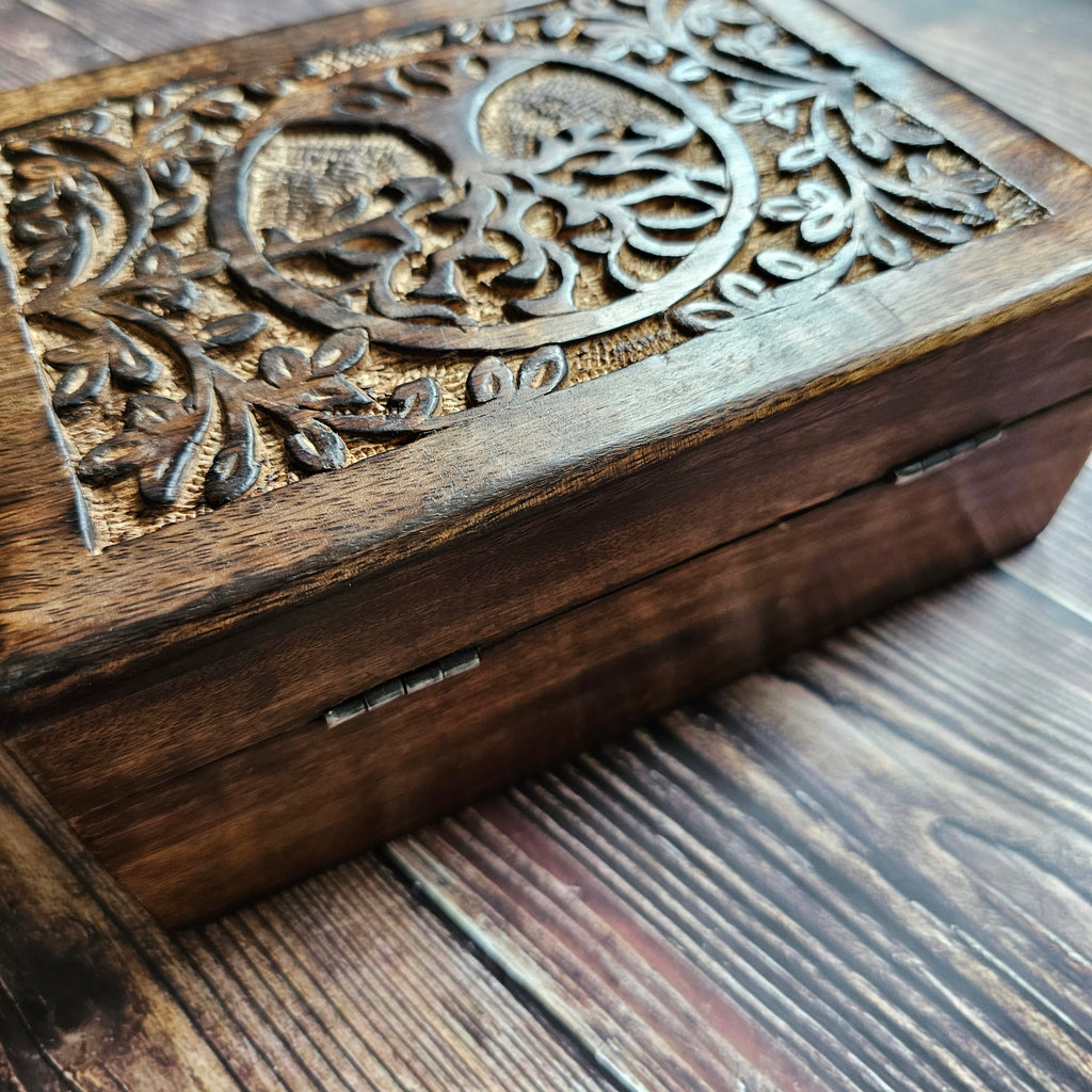 A wooden box with a carved Tree of Life design on the lid, placed on a wooden surface.