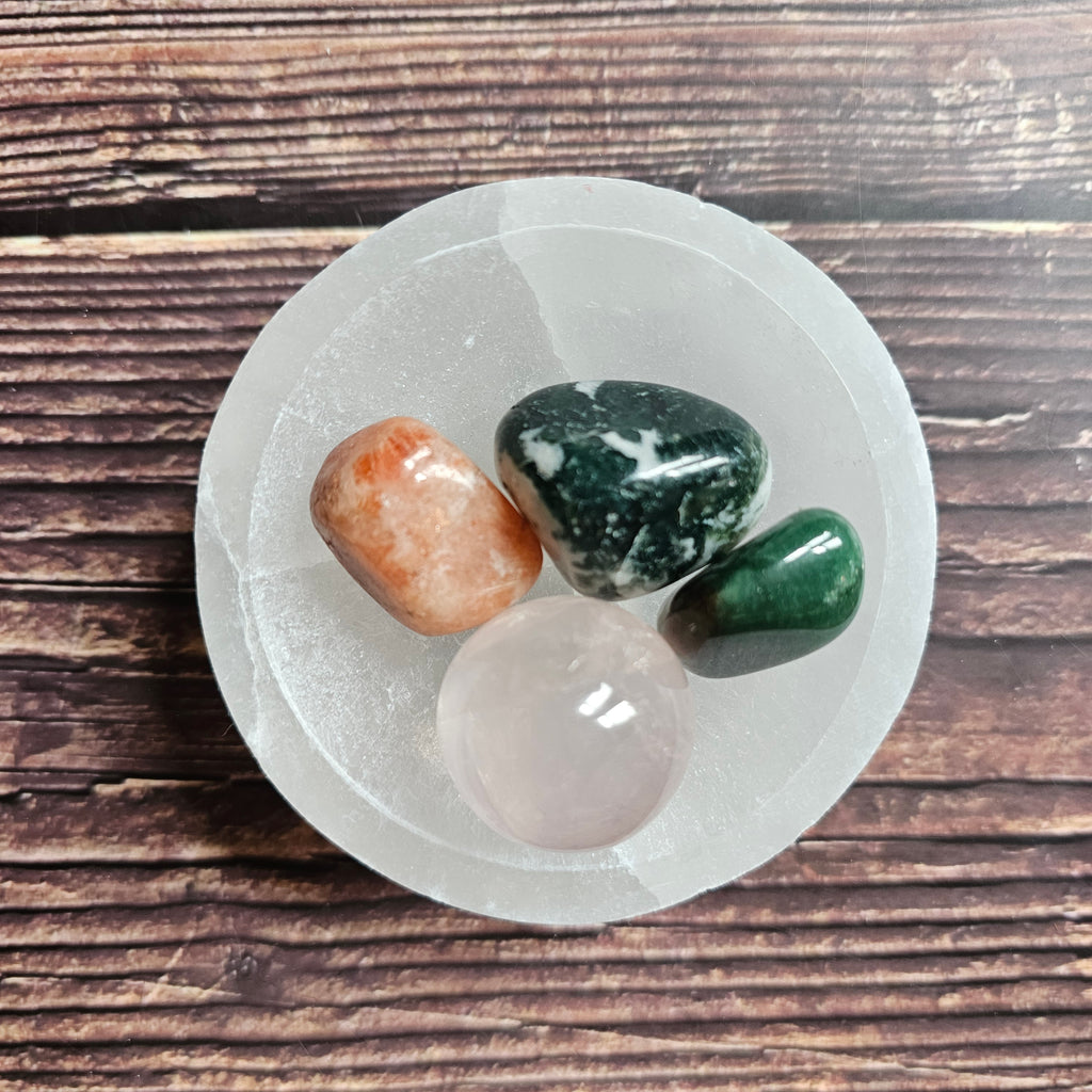 A white selenite bowl on a wooden surface, with several small colorful stones arranged on it.