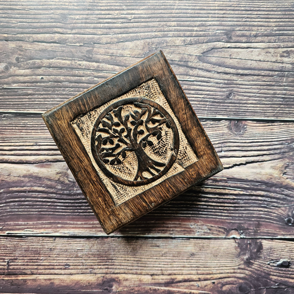 A square wooden box with a carved Tree of Life design on the lid, placed on a wooden surface.