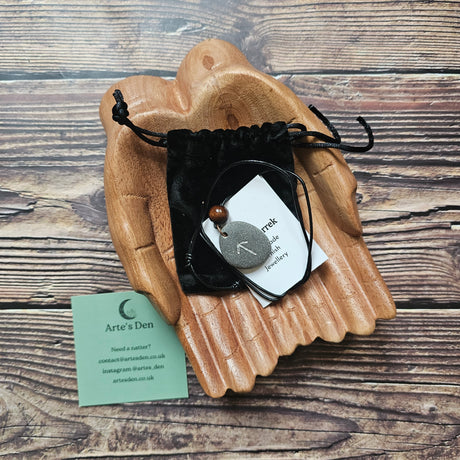 Hand-carved rune stone necklace, featuring Tiwaz rune, on a black cord. Displayed on a wooden pair of hands, sat on a wooden surface next to a business card. 