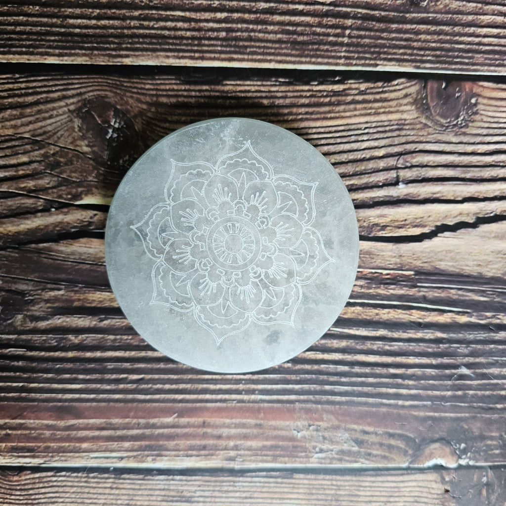 A round selenite charging plate with a mandala pattern etched into it, placed on a wooden surface.