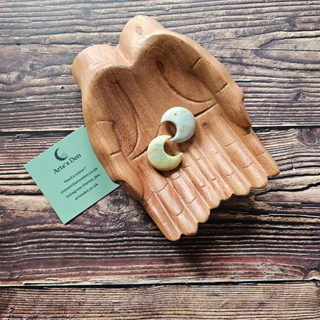 Two amazonite crescent moon shaped crystals placed on a pair of wooden hands on a wooden surface, with a business card labelled 'Arte's Den'.