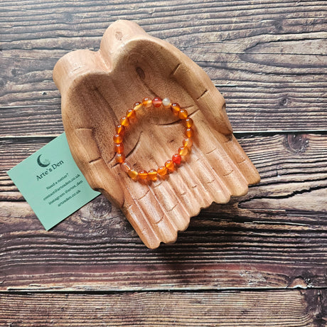 A translucent deep orangey-red carnelian beaded bracelet displayed on a wooden hand-shaped tray with a wooden background.