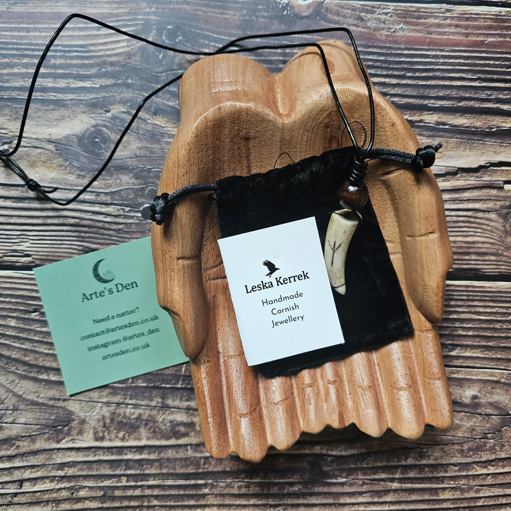 A handcarved antler pendant necklace with a black cord, featuring the Algiz Rune, placed on a wooden surface with a business card beside it.