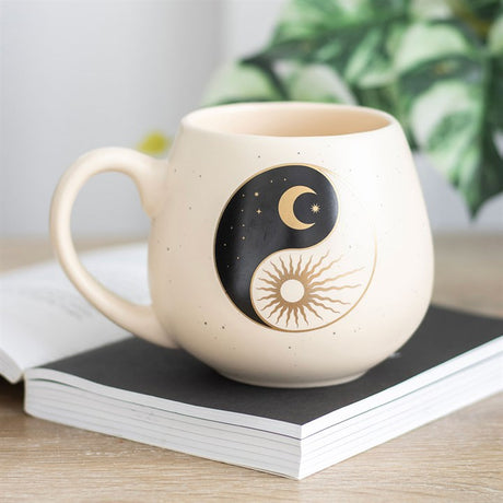 A beige bone china mug with a black and gold yin and yang design, featuring a sun and crescent moon symbol, placed on a wooden surface next to a plant and a book.
