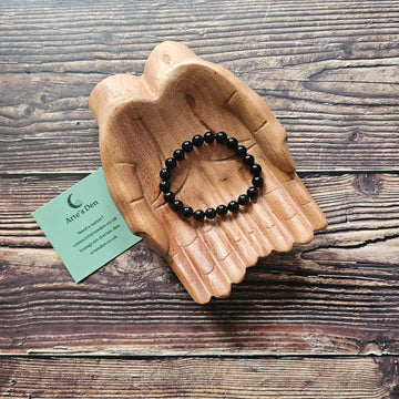 A black obsidian beaded bracelet displayed on a wooden pair of hands with a business card from Arte's Den.