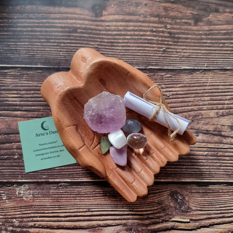 A collection of crystals and a scroll of paper displayed in a pair of wooden hands, with a business card that reads 'Arte's Den'.