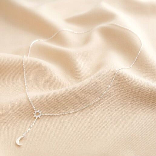 A silver lariat necklace featuring a crescent moon and an open sun charm laid on a light background. 