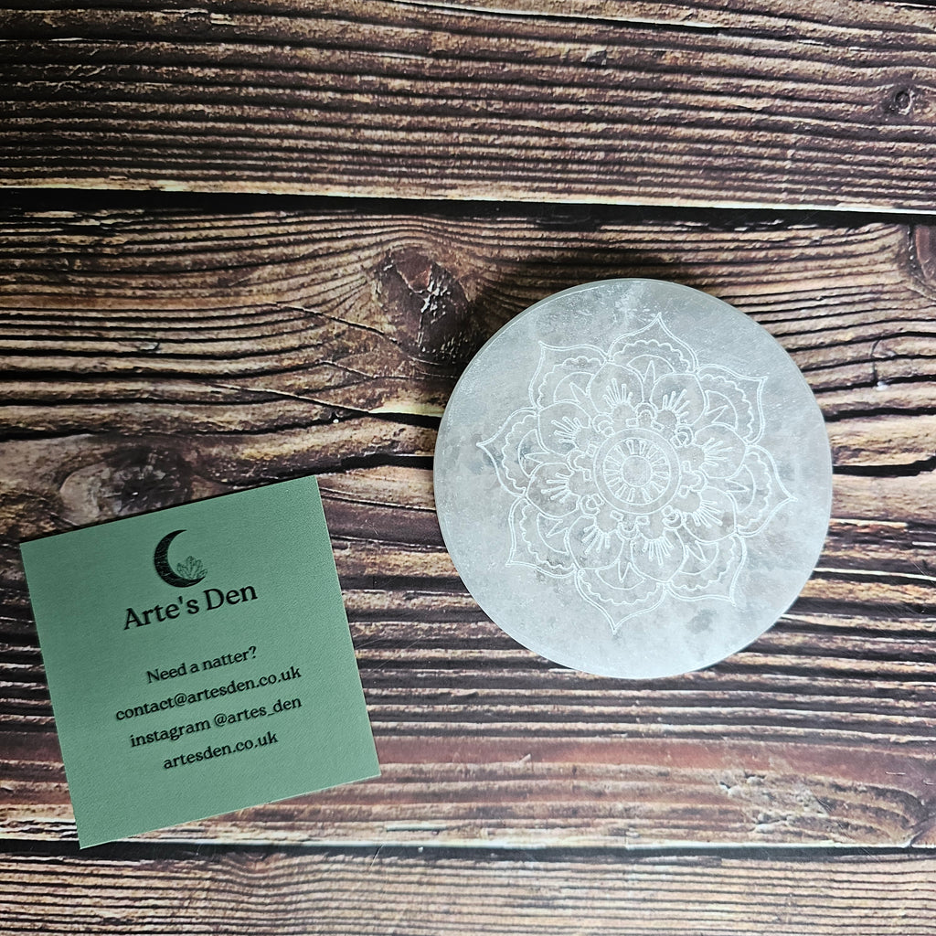 A round selenite charging plate with a mandala pattern etched into it, placed on a wooden surface.