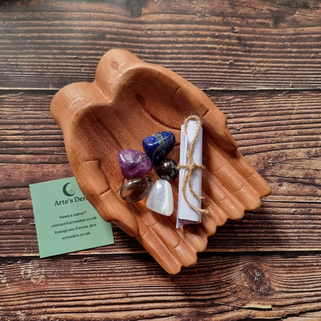 A collection of polished crystals and a scroll of paper displayed in a pair of wooden hands, with a business card that reads 'Arte's Den'.